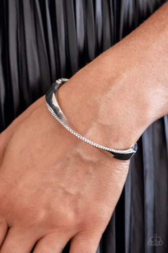 An oversized, warped, shiny silver cuff wraps around the wrist in an artistic manner. A dainty row of white rhinestones encrusts along the curves of the cuff for an understated, elegant dazzle. Features a hinged closure.  Sold as one individual bracelet.