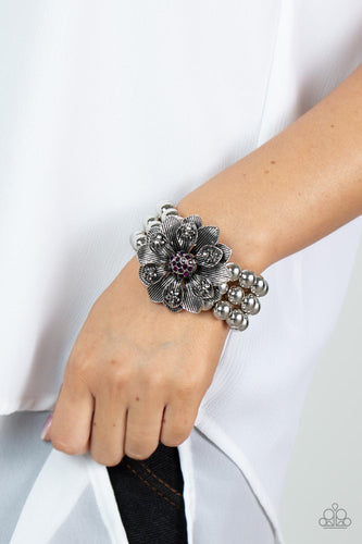 A daring oversized silver flower is composed of petals lined in antiqued silver and dotted with smoky hematite rhinestones. A sphere of dainty purple rhinestones creates the center of the flower as it sits atop a trio of shiny silver beaded stretchy bands around the wrist for a dramatically whimsical look.  Sold as one individual bracelet.