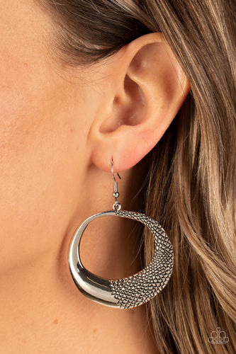 One side of an asymmetrical silver hoop is embossed in rugged pebble like texture, culminating in a wildly abstract frame. Earring attaches to a standard fishhook fitting.  Sold as one pair of earrings.